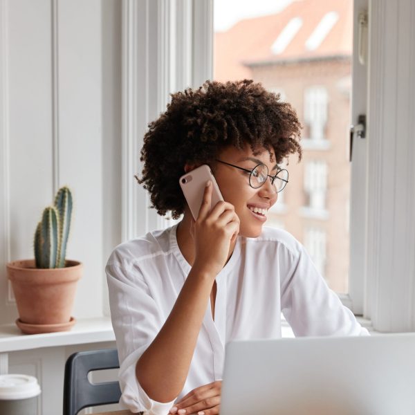Vertical shot of cheerful woman bookkeeper talks on mobile phone, has positive expression, dressed in white stylish blouse, works with laptop computer, has work break, drinks hot aromatic beverage